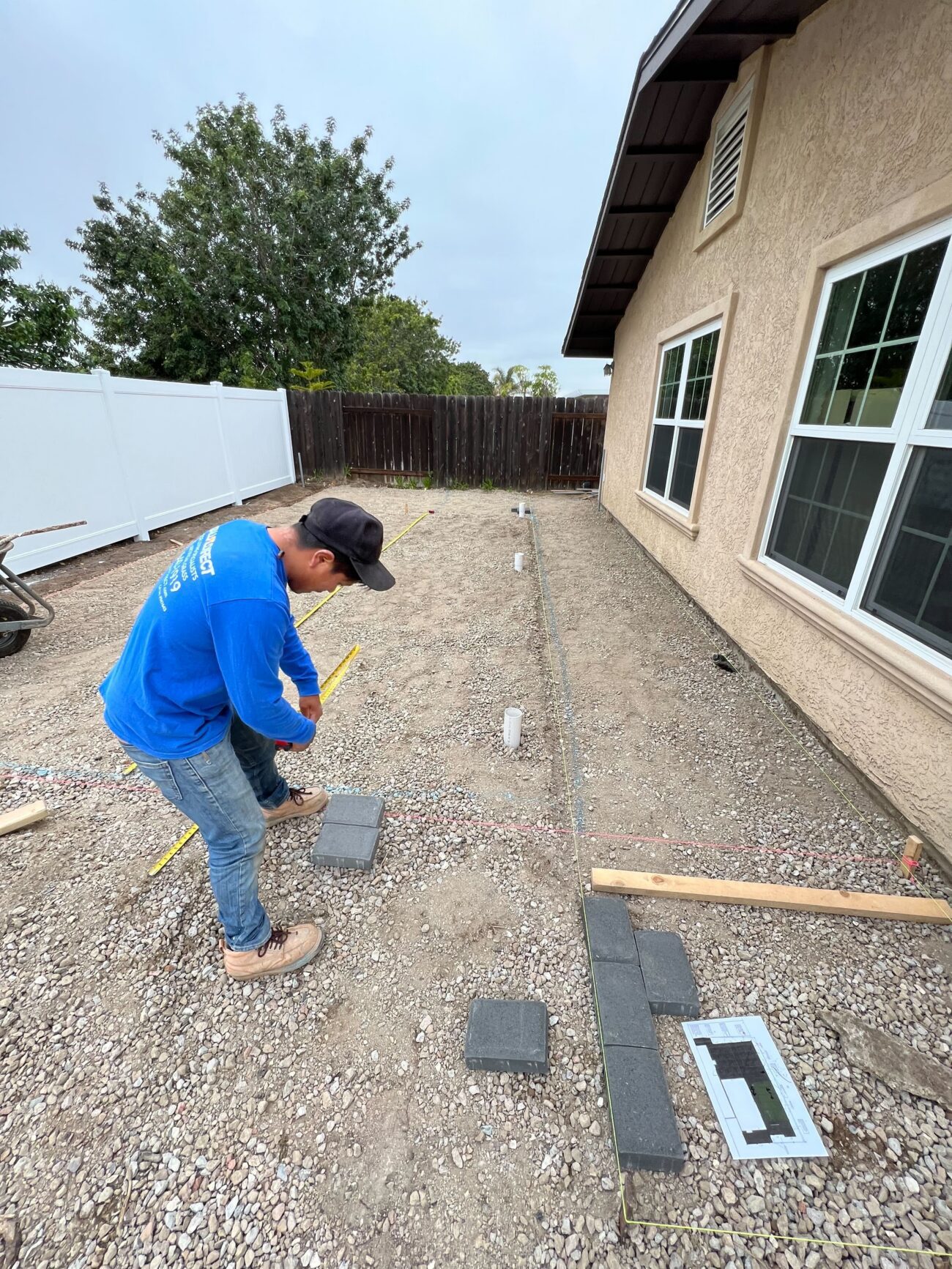 A crew member is measuring and marking layout on a compacted gravel base adjacent to a house, with string lines and PVC...