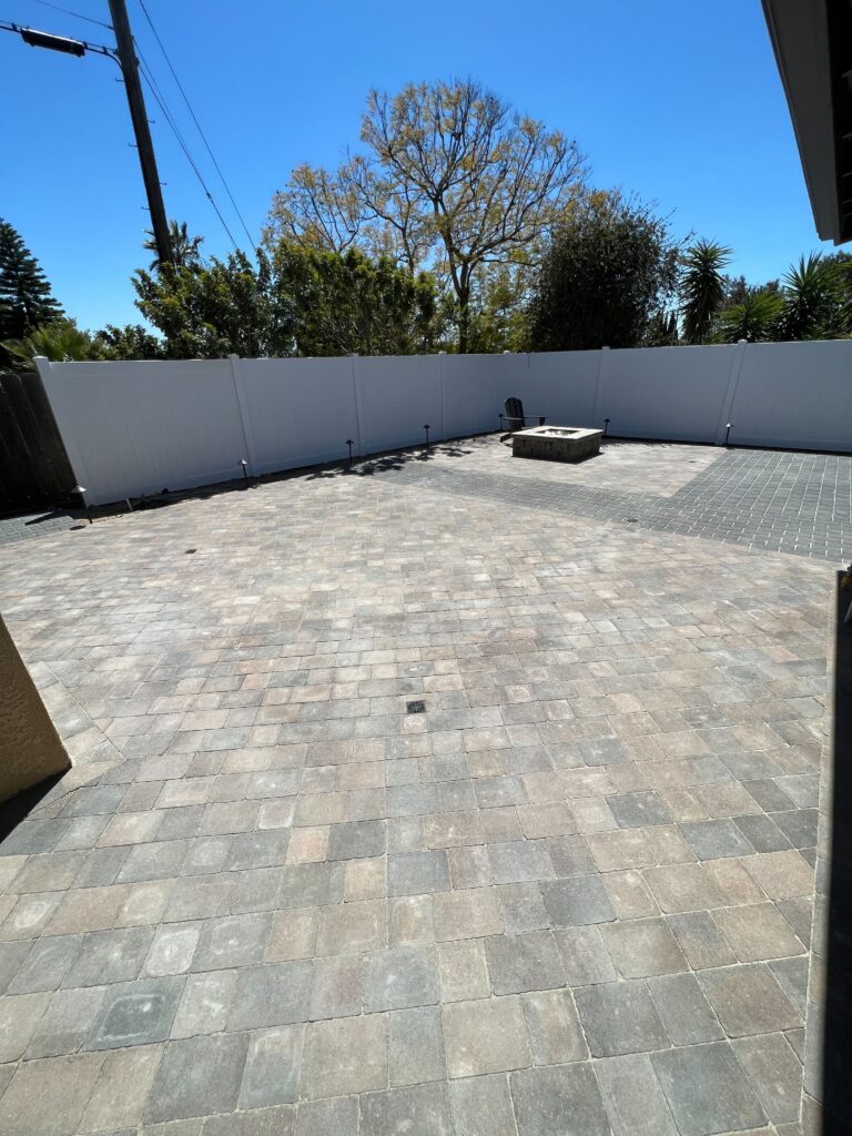 Finished interlocking paver patio in a residential backyard with a centrally located square fire pit and a vinyl privacy...
