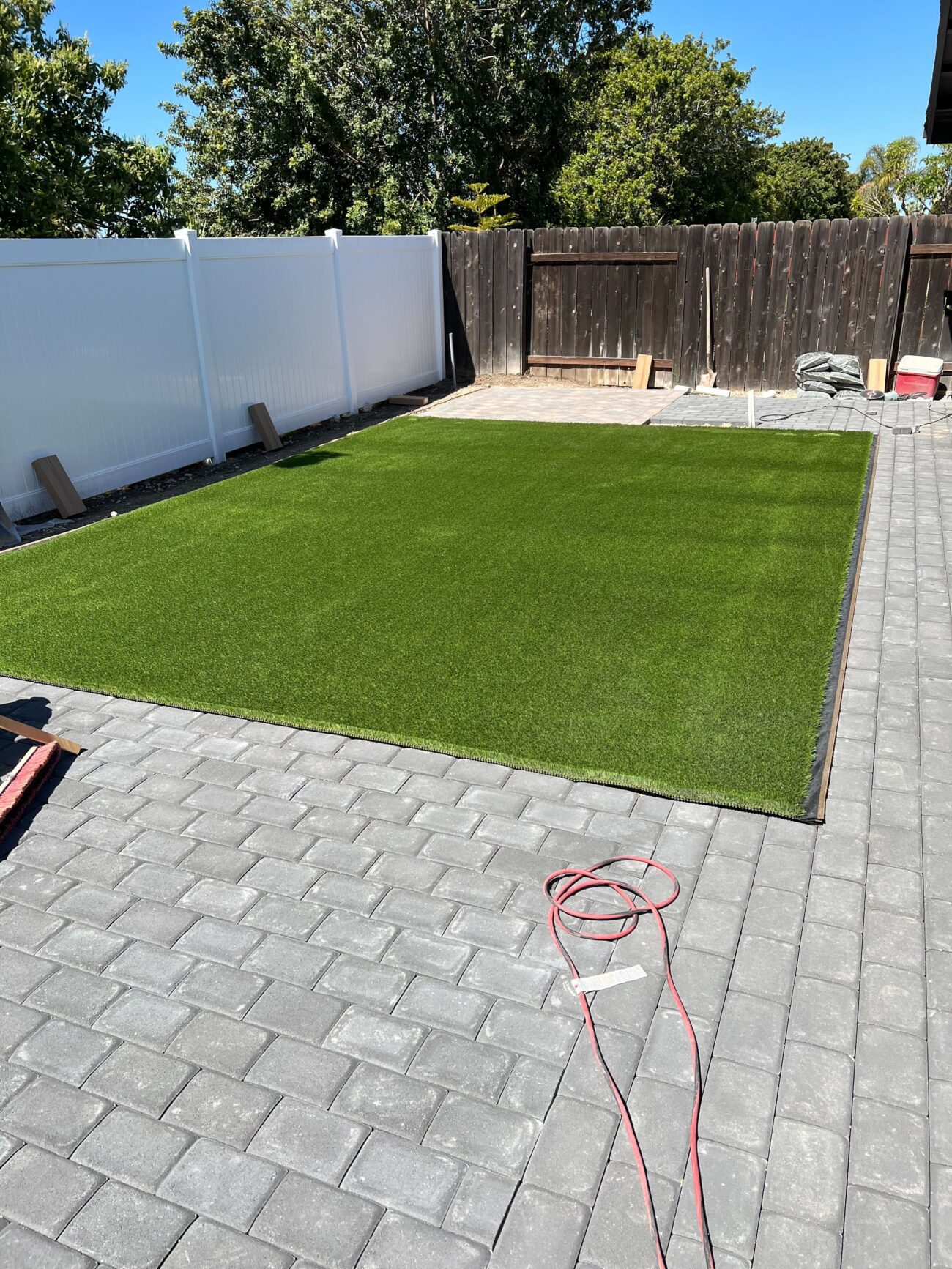 Rectangular artificial turf pad has been installed flush within a field of interlocking gray pavers in a residential...
