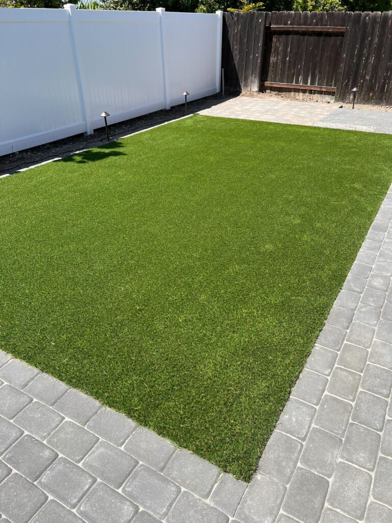 Finished backyard installation showing a rectangular artificial turf lawn bordered by gray interlocking pavers. White...