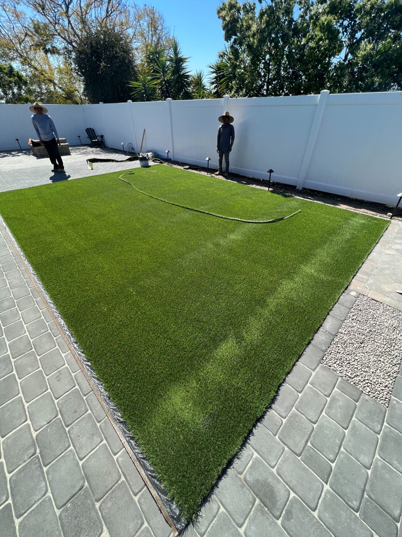 Residential backyard showing a newly laid artificial turf panel bordered by interlocking pavers and a small gravel strip....