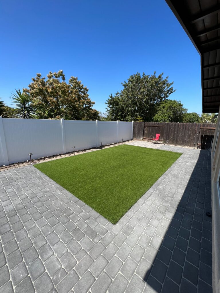 Backyard showing a completed artificial turf rectangle surrounded by interlocking pavers forming a patio and walkway....