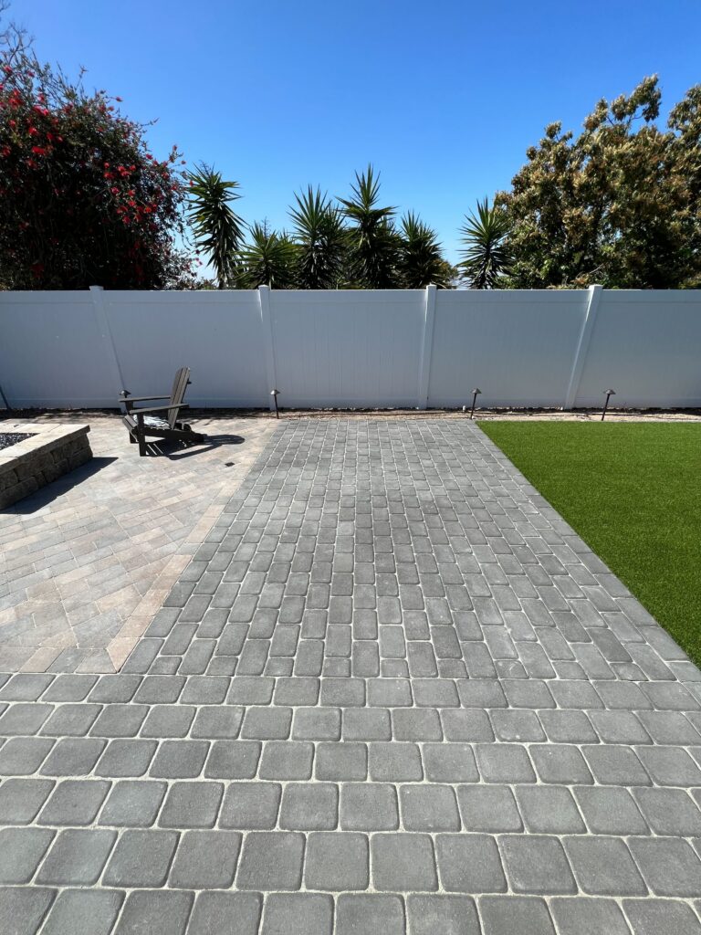 Finished backyard showing a gray interlocking paver patio transitioning to installed artificial turf with a low segmental...