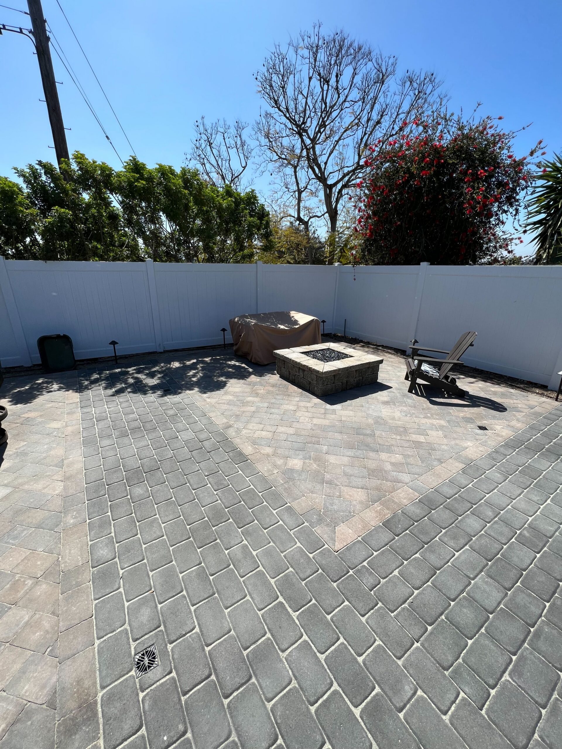 Completed interlocking paver patio with a central square stone-veneer fire pit and contrasting inset field pattern. White...