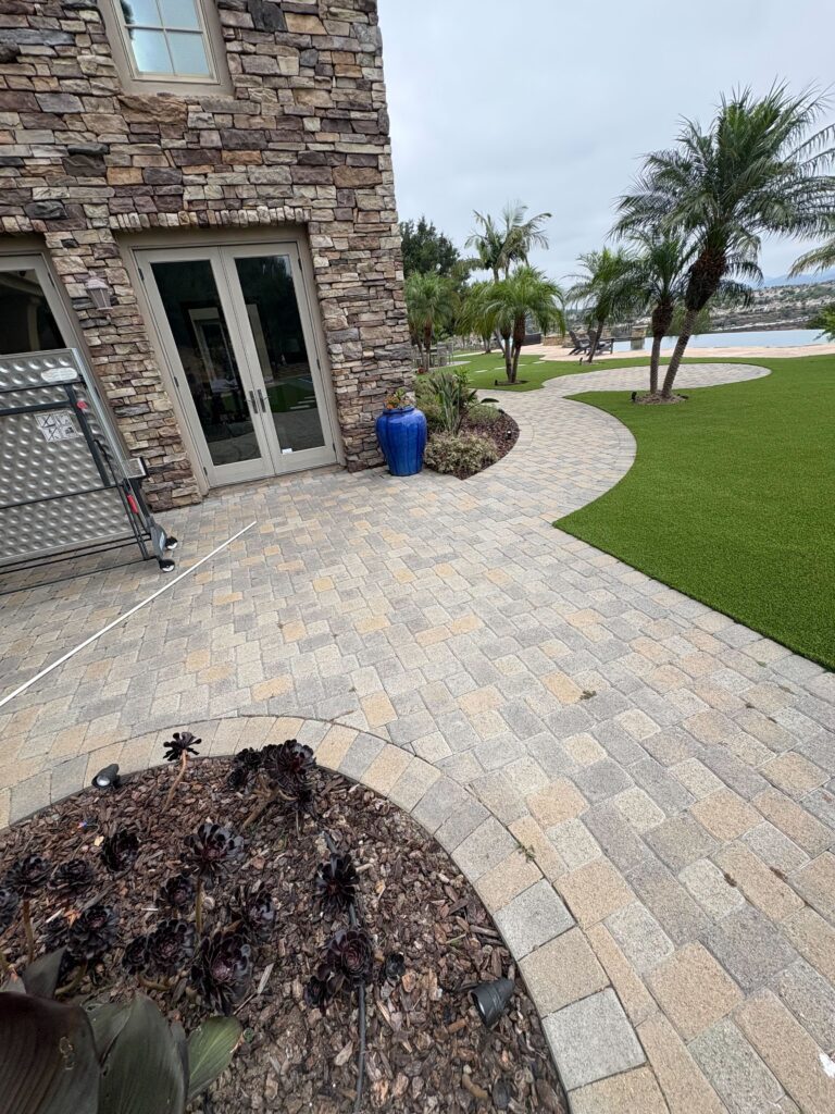 Finished backyard showing a curved interlocking paver patio and walkway adjacent to installed artificial turf. Stone...