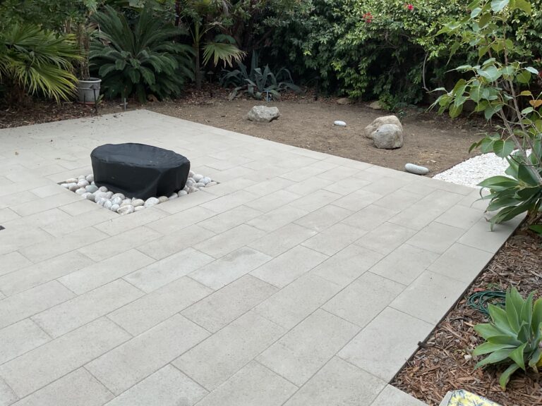 A recently installed light-gray rectangular paver patio in a backyard with a covered fire pit set in a small bed of river...