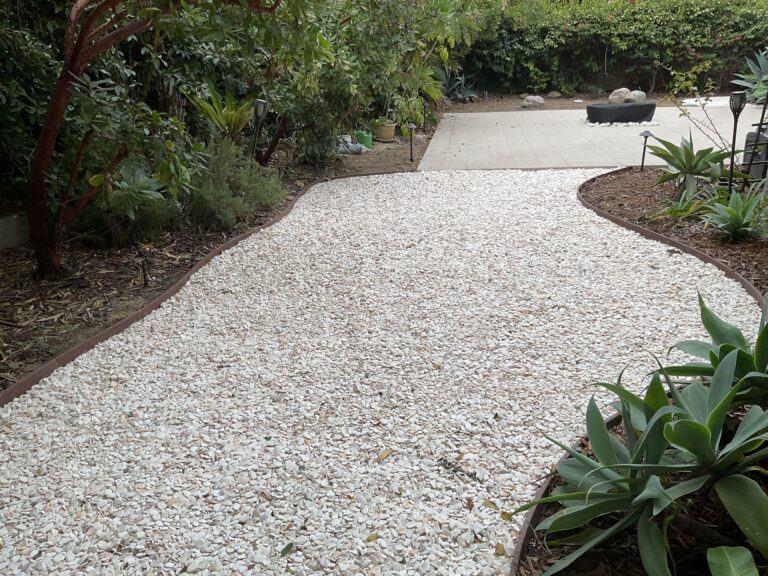 Finished backyard pathway surfaced with light crushed stone edged by metal/band edging, flanked by planting beds and...