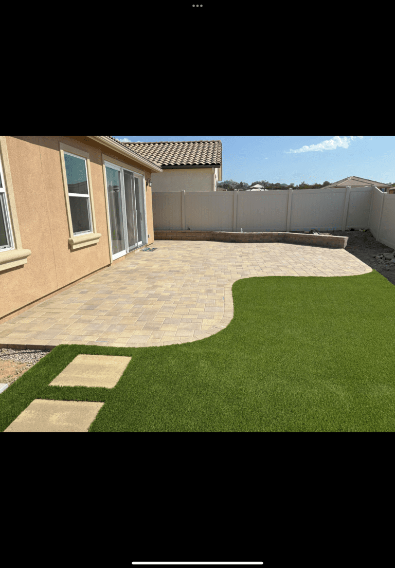 Newly installed interlocking paver patio with clean swept joints abutting a neatly trimmed artificial turf lawn and...