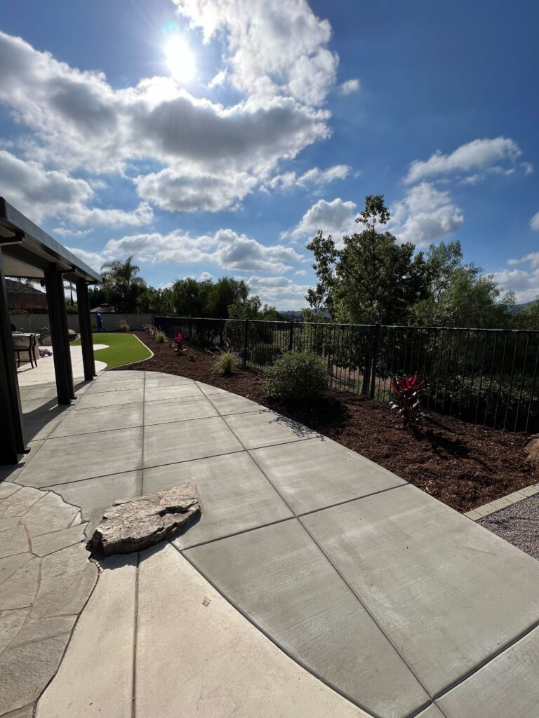 Sunlit backyard showing a newly finished poured concrete patio and curved walkway with saw-cut control joints and a broom...