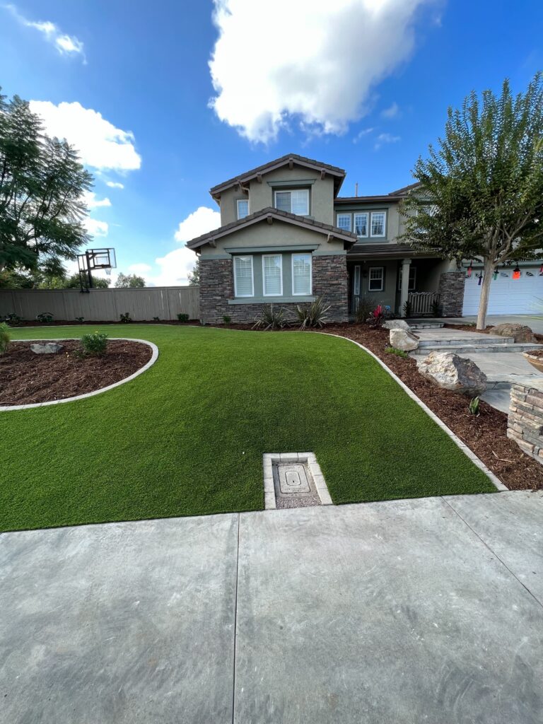Front yard of a two-story residence showing newly installed artificial turf with a concrete driveway, curved mulch...