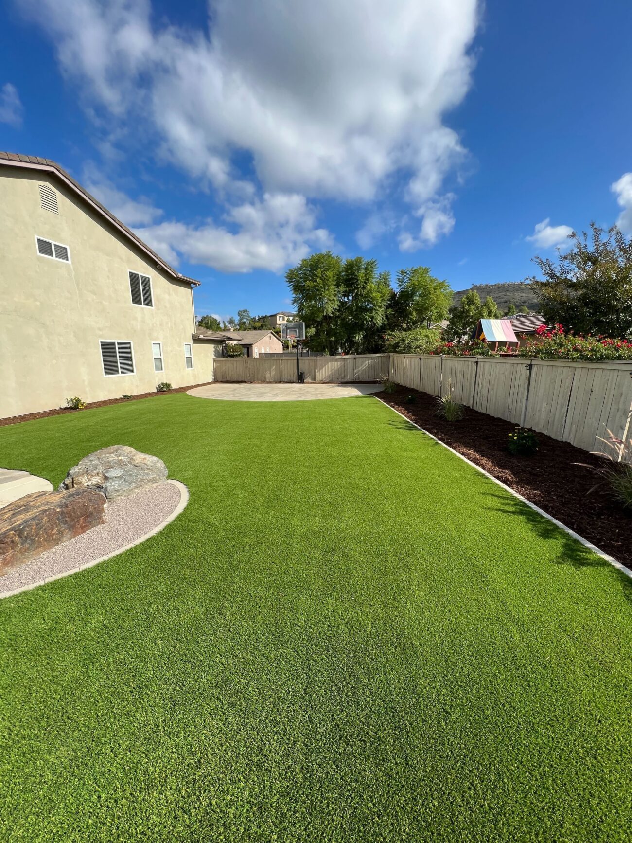 Installed artificial turf covers a suburban backyard with a concrete patio slab at the far end and decorative boulders...