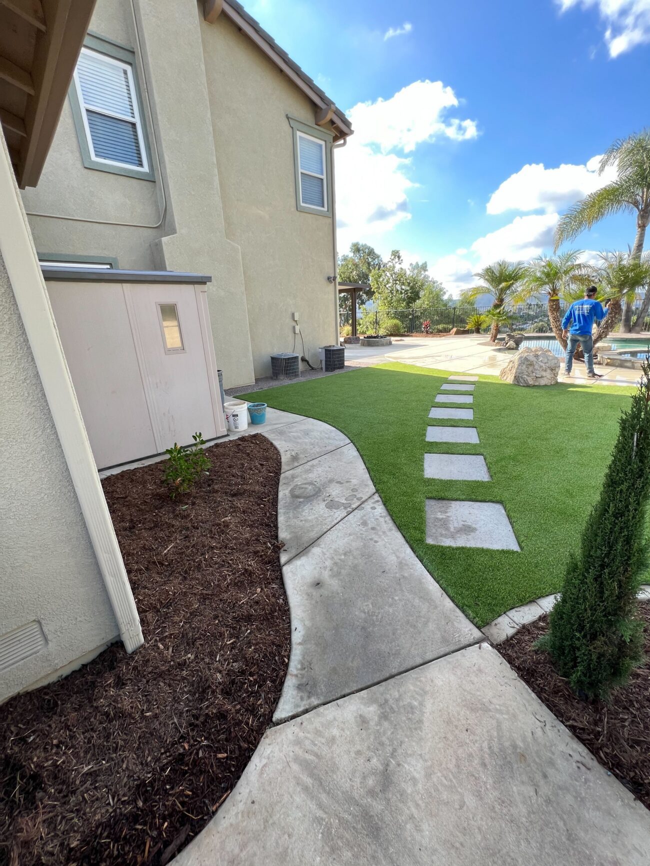 Backyard showing newly installed artificial turf with square paver stepping stones integrated into the turf and a curved...
