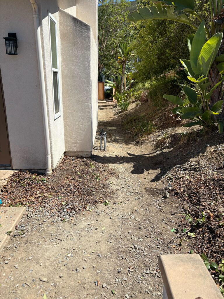 Narrow residential side yard with a compacted dirt access path running alongside a stucco exterior wall and down a...