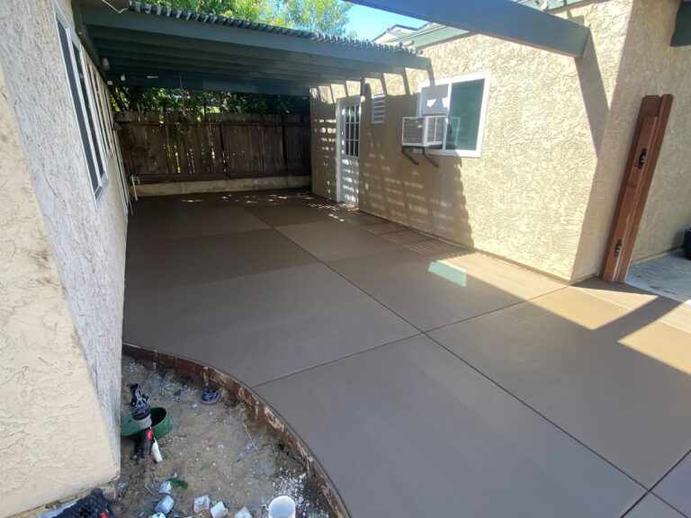 Residential backyard showing a newly finished colored concrete patio with saw-cut control joints under a covered pergola....