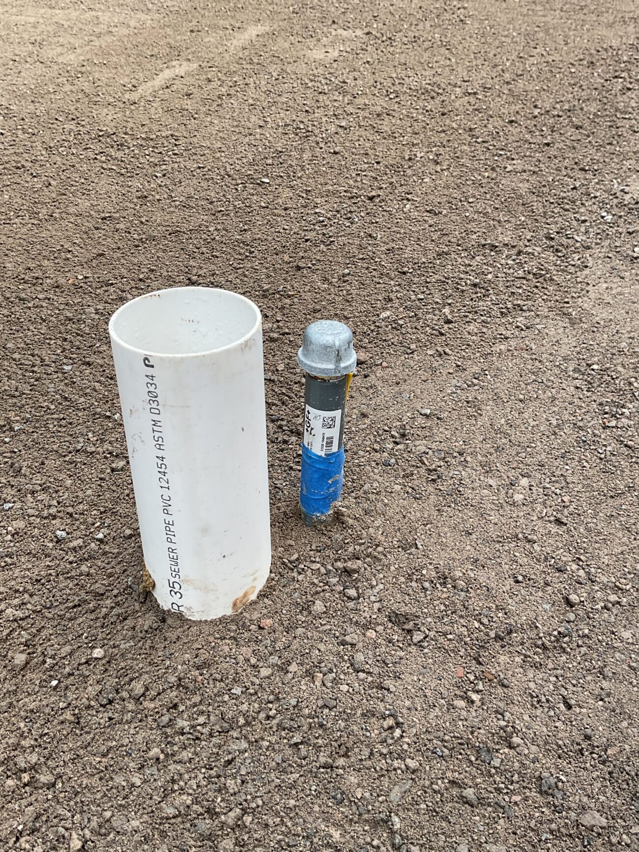 Compacted aggregate base with two utility stubs protruding through the subbase: a larger PVC riser (likely a cleanout or drainage stub) and a smaller capped service stub wrapped in blue tape. San Diego project by Install It Direct.