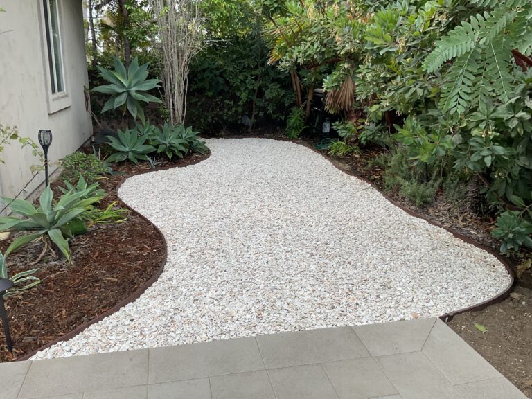 Finished side-yard landscape showing a curved field of decorative white gravel/river rock with black landscape edging, adjacent to a small paver patio. San Diego project by Install It Direct.
