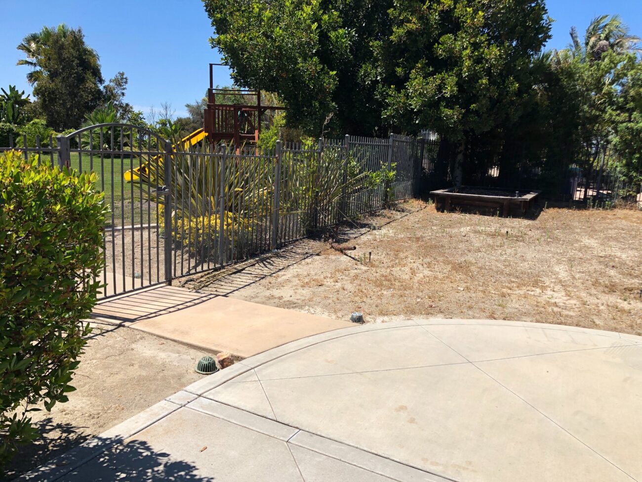 Concrete patio slab with a curved edge sits adjacent to a compacted dirt yard and a metal picket fence enclosing a play...