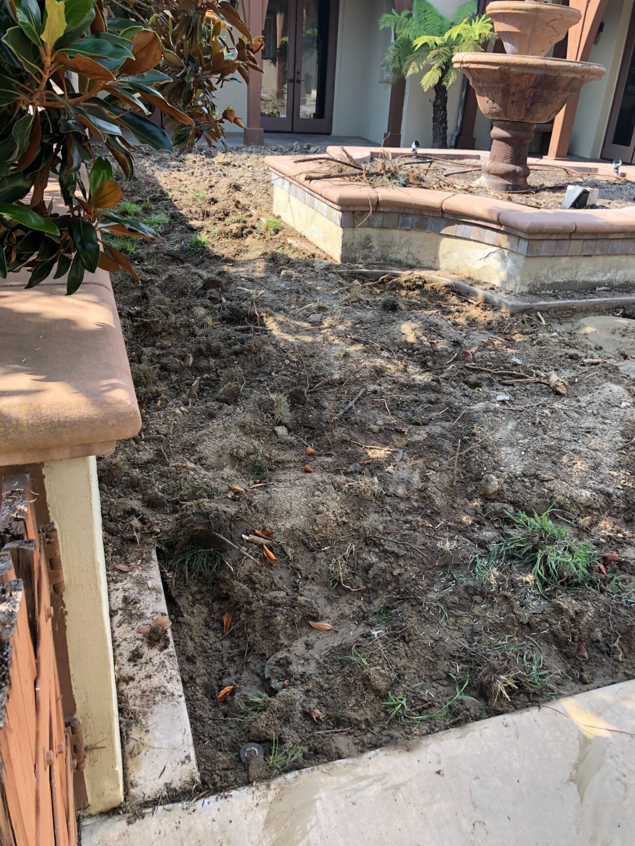 Backyard landscape area with turf removed and surface excavated adjacent to a raised stone planter and fountain. Soil is...