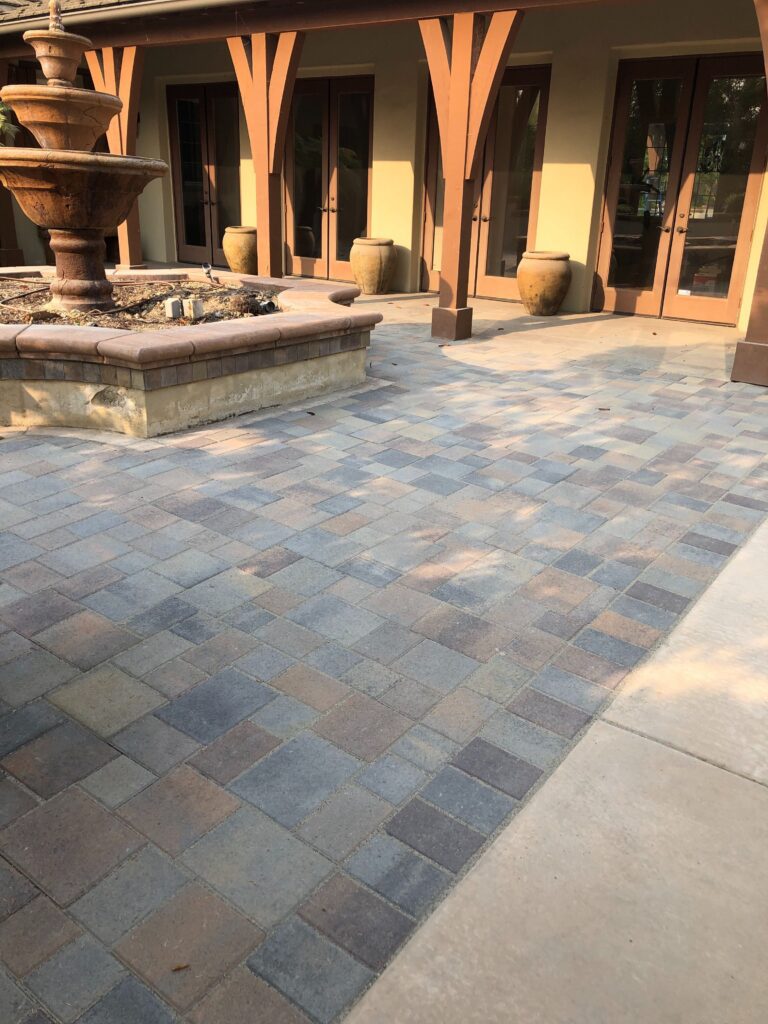Completed interlocking paver patio adjacent to a residential exterior with a raised stone planter and fountain. Concrete...