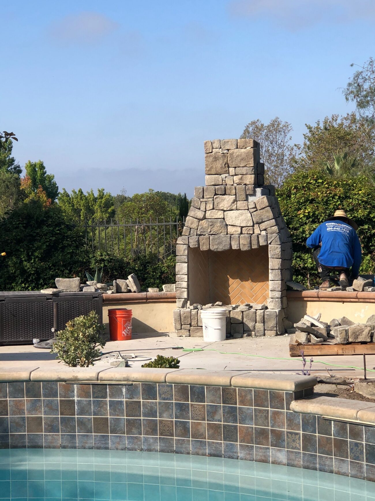 Stone outdoor fireplace under construction on a tiled pool deck with adjacent paver patio. Crew member is on-site with...