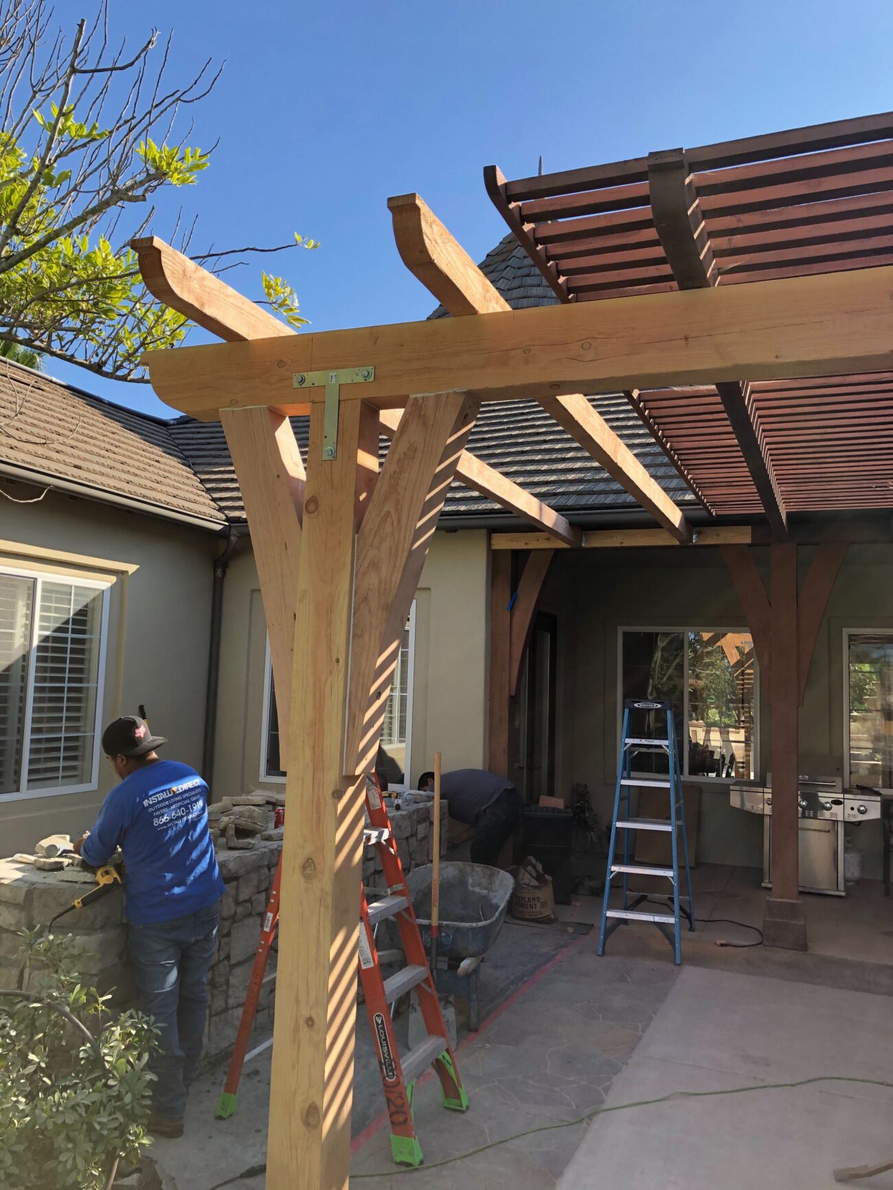 Timber post-and-beam pergola being installed over a backyard patio; large wooden posts and rafters are in place with...