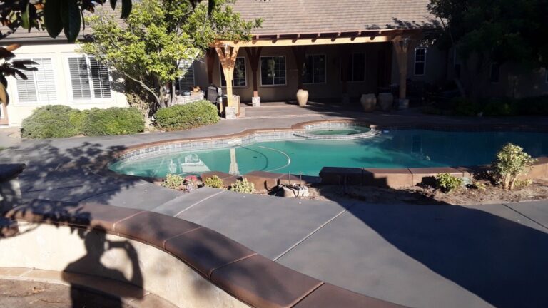 Residential backyard showing a completed pool deck and adjacent covered patio. Broom-finished concrete deck with control...