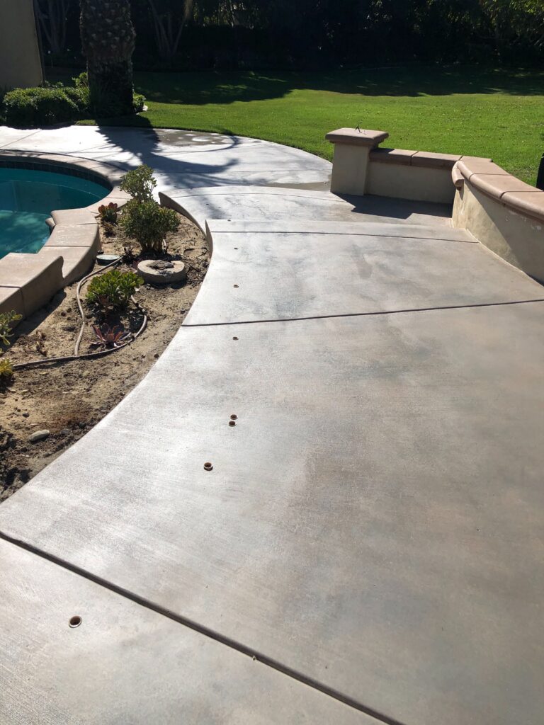 Finished concrete pool deck with visible control/expansion joints and several embedded anchor plugs. The deck ties into a...