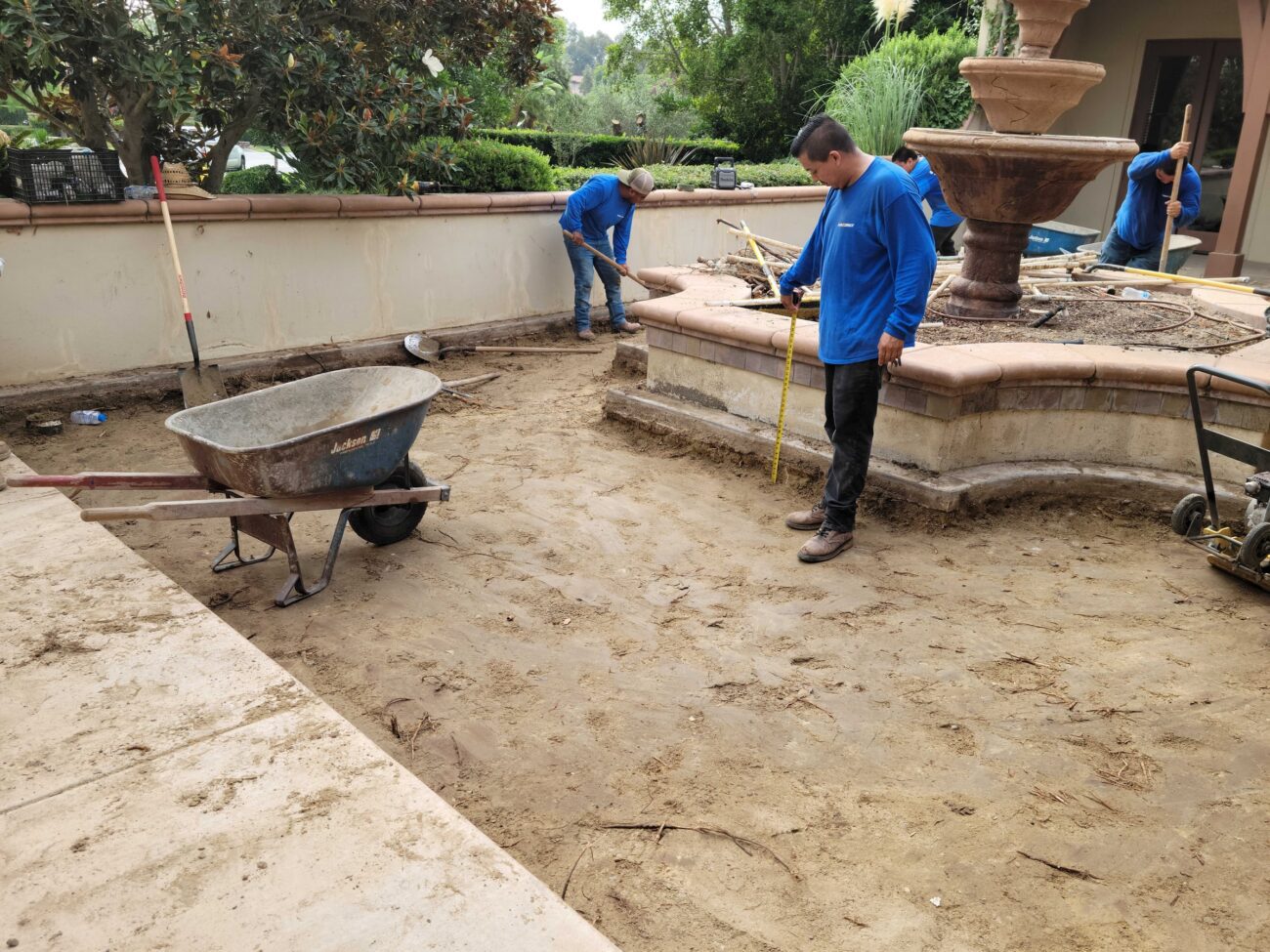 Crew members are preparing the subgrade of a residential patio surrounding a multi-tiered stone fountain and existing low...