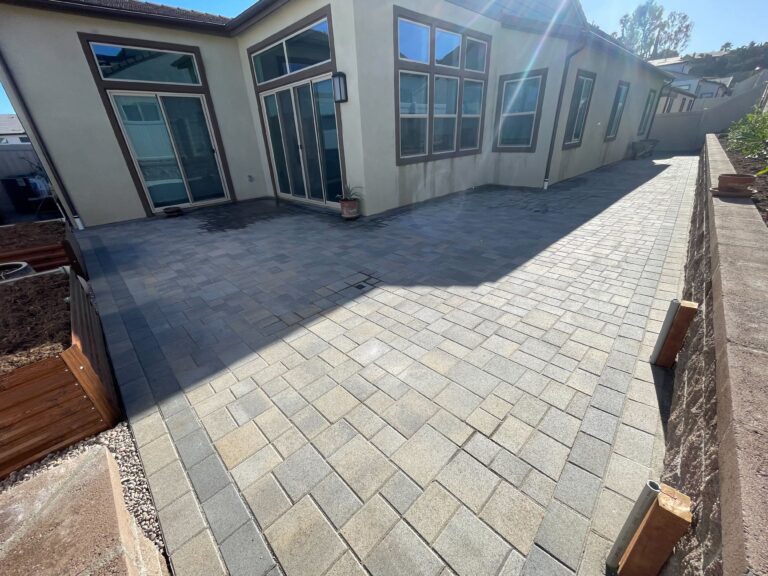 Finished interlocking concrete pavers forming a large patio/walkway adjacent to a stucco residential exterior. Modular...
