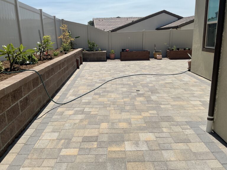 Completed interlocking paver patio in a residential backyard with a segmental block retaining/planter wall along the left...