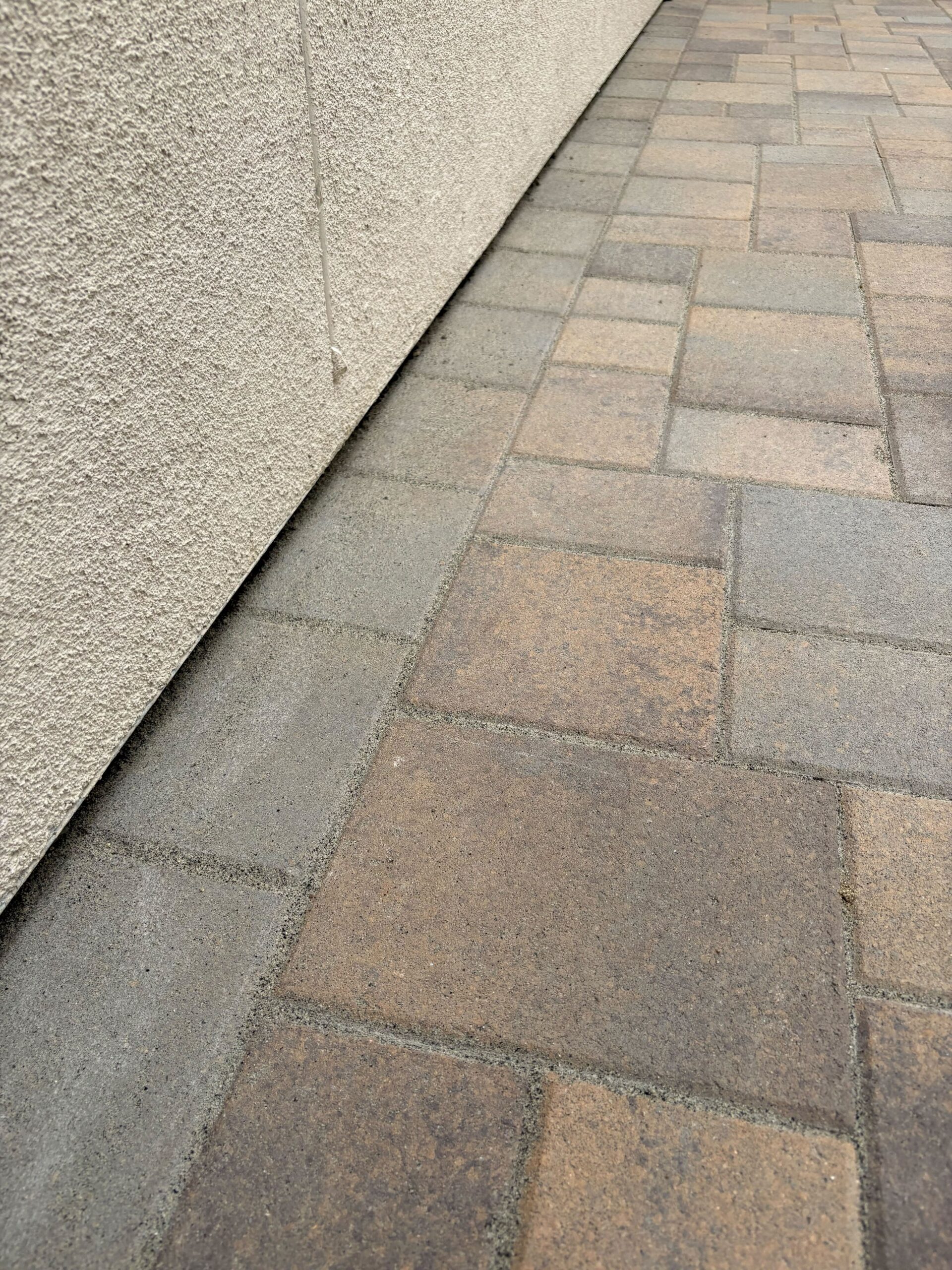 Close-up photo of a newly installed interlocking paver walkway running alongside a stucco exterior wall. Mixed-size...