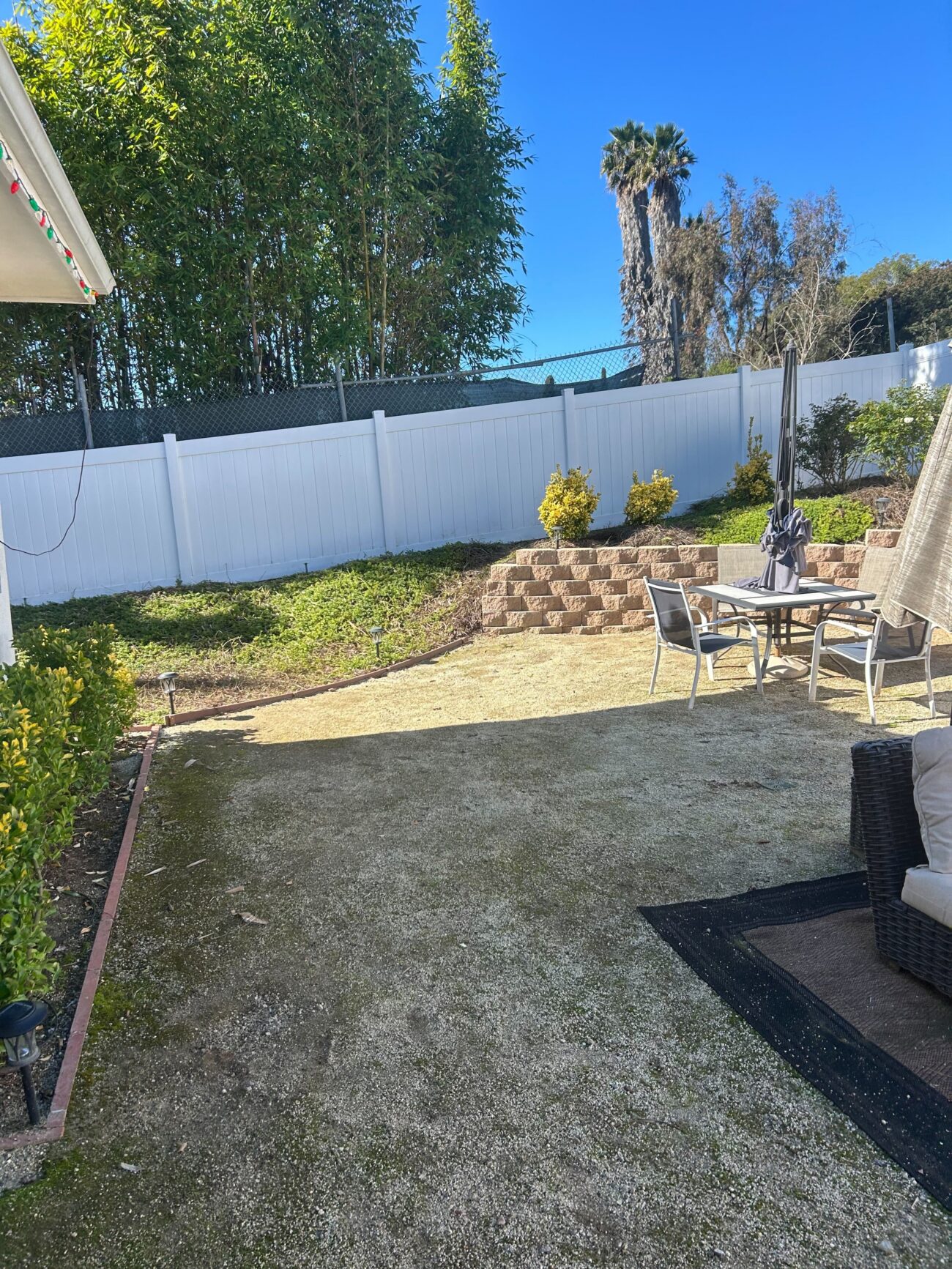 Vista backyard before renovation: patchy natural turf and bare soil sloping toward a segmental retaining wall and white vinyl fence. Install It Direct project.