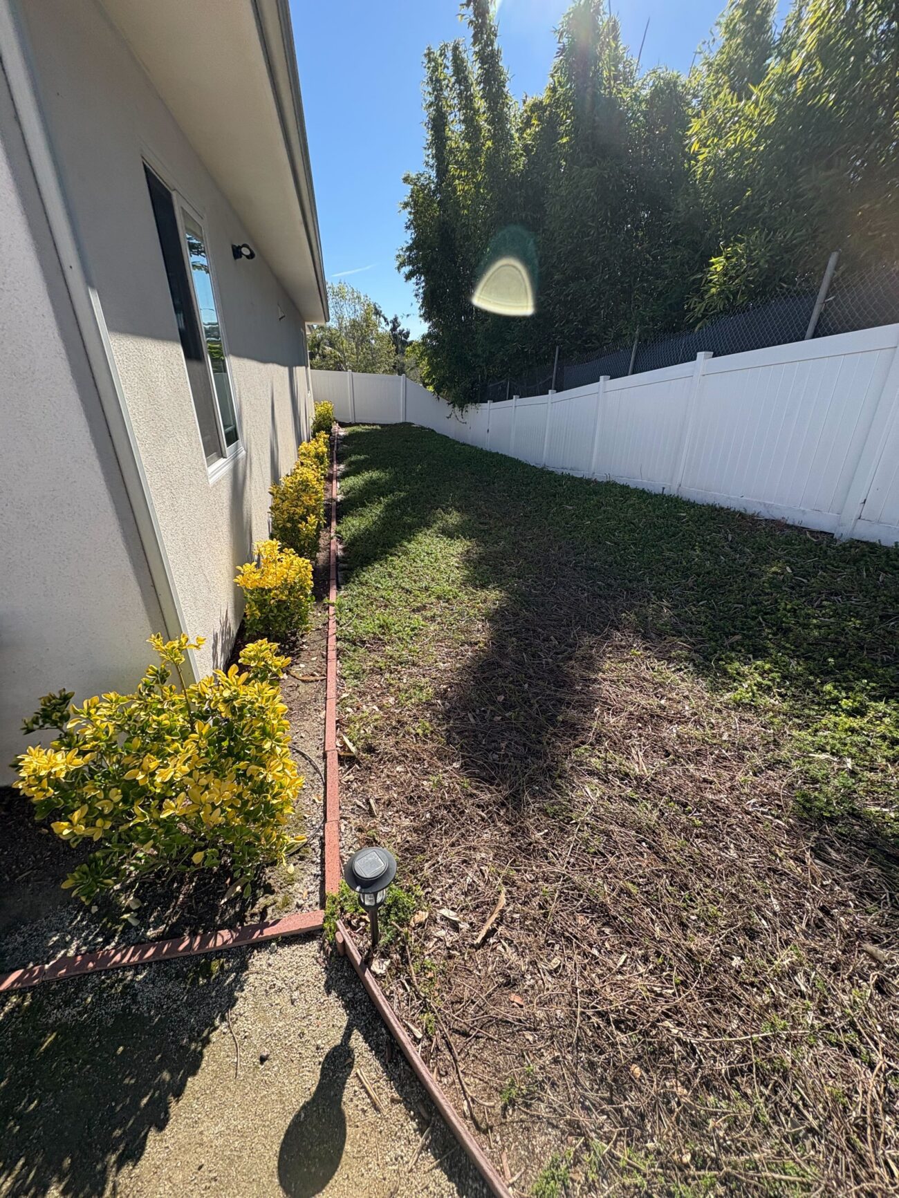 Vista side yard before renovation: narrow corridor with patchy turf, exposed soil, and brick edging beside the stucco house and vinyl fence. Install It Direct project.