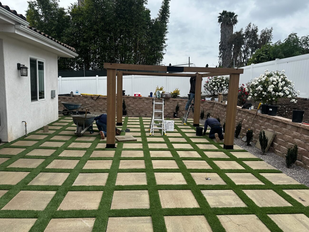 Backyard during active installation: paver-and-turf grid with crew assembling a wooden pergola against the perimeter retaining wall in Vista, CA. Install It Direct project.