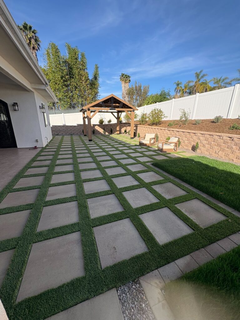 Checkerboard layout of large concrete pavers with artificial-turf joints, pergola, and segmental retaining wall in a Vista backyard. Install It Direct project.