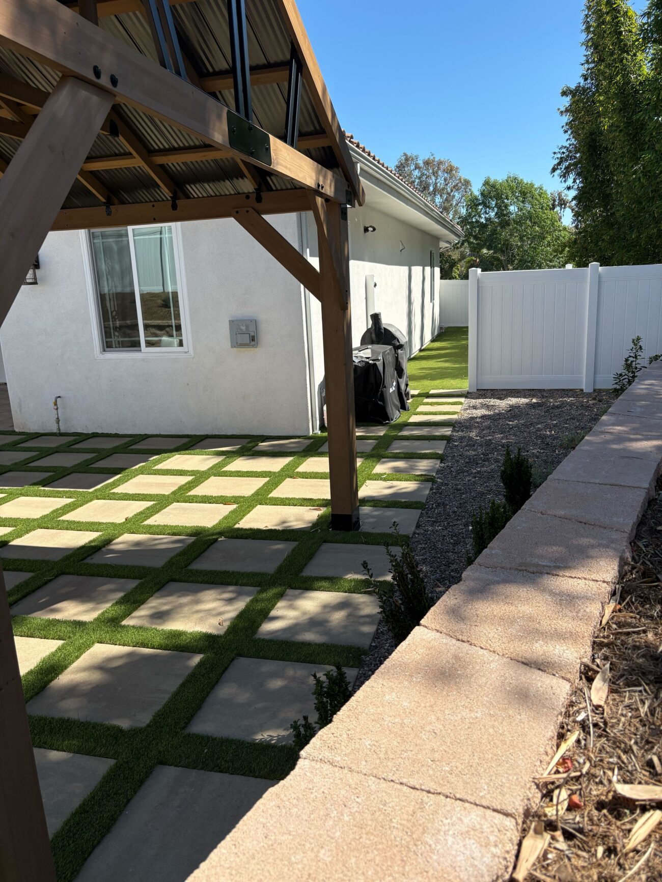 Timber pergola over paver-and-turf grid with low retaining wall and decorative stone border along the side yard in Vista, CA. Install It Direct project.