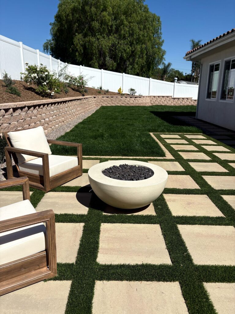 Bowl fire feature and seating on the paver-and-turf grid against the segmental retaining wall and white vinyl fence in Vista, CA. Install It Direct project.