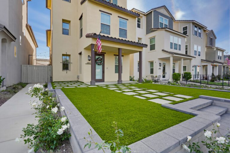 Front yard of a townhouse showing a completed artificial turf lawn with large concrete paver stepping stones set in a grid pattern and a low segmental retaining wall forming raised planting beds. Escondido project by Install It Direct.