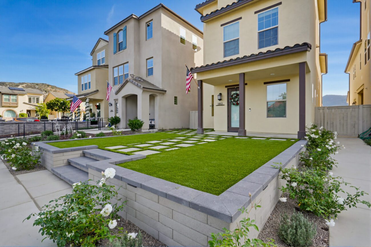 Front yard of a single-family home showing finished artificial turf with large square paver stepping stones set flush in the turf adjacent to a concrete walkway. Escondido project by Install It Direct.