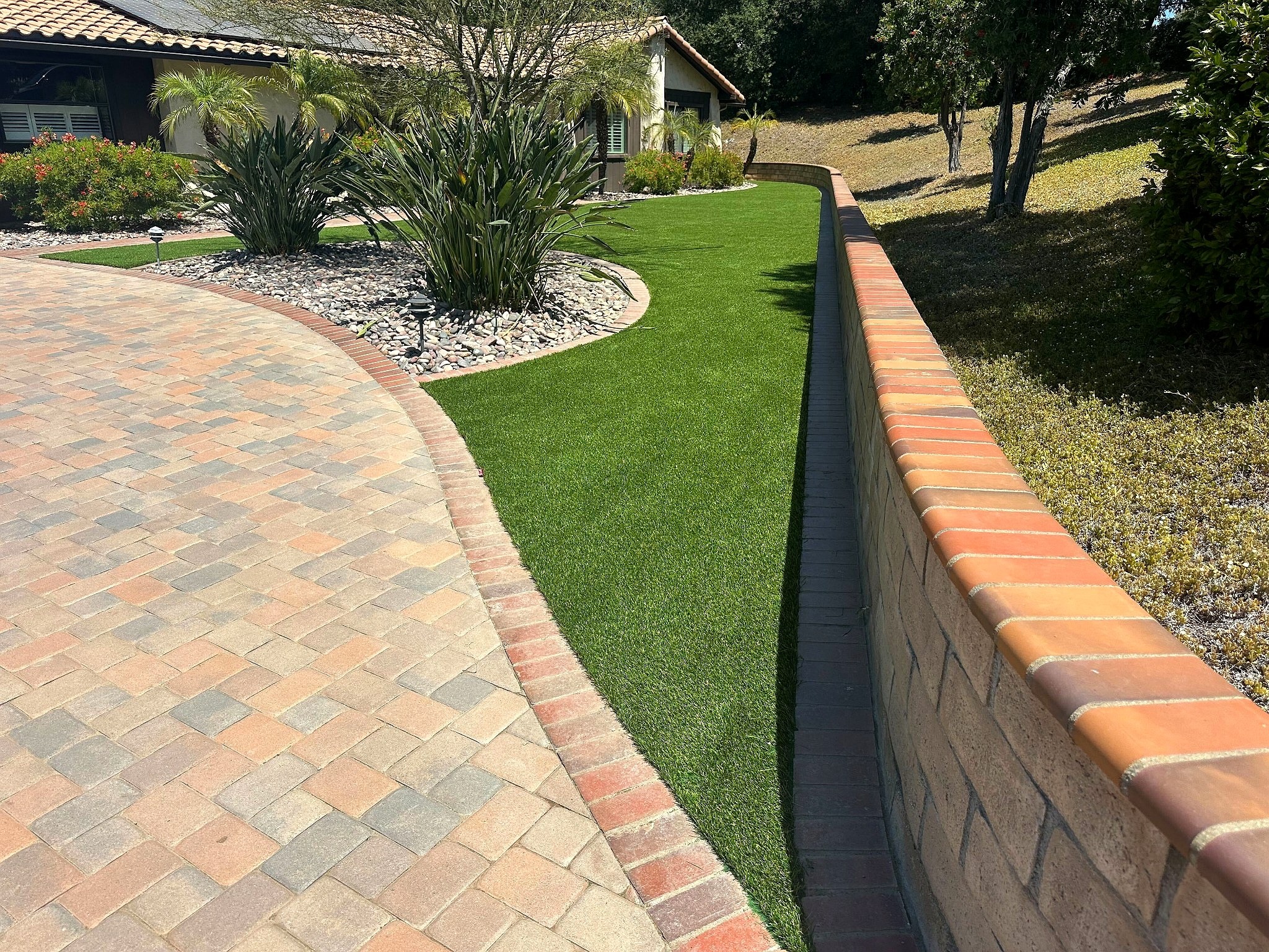 SiteOne Rye turf strip between the paver driveway and a terracotta-capped retaining wall, with river-rock planting beds at the house.