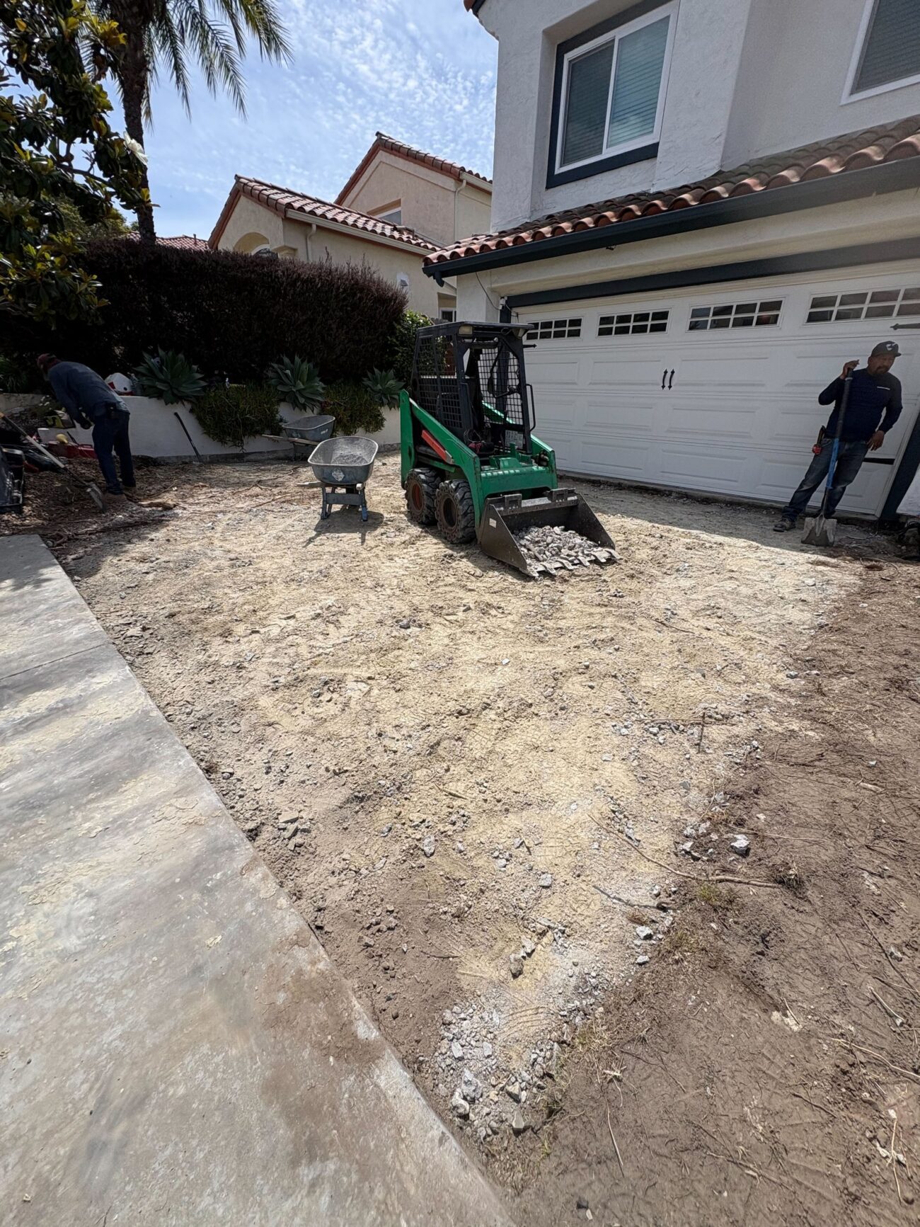 Residential driveway demolition in progress: a skid-steer is removing broken concrete and rubble from the driveway area...