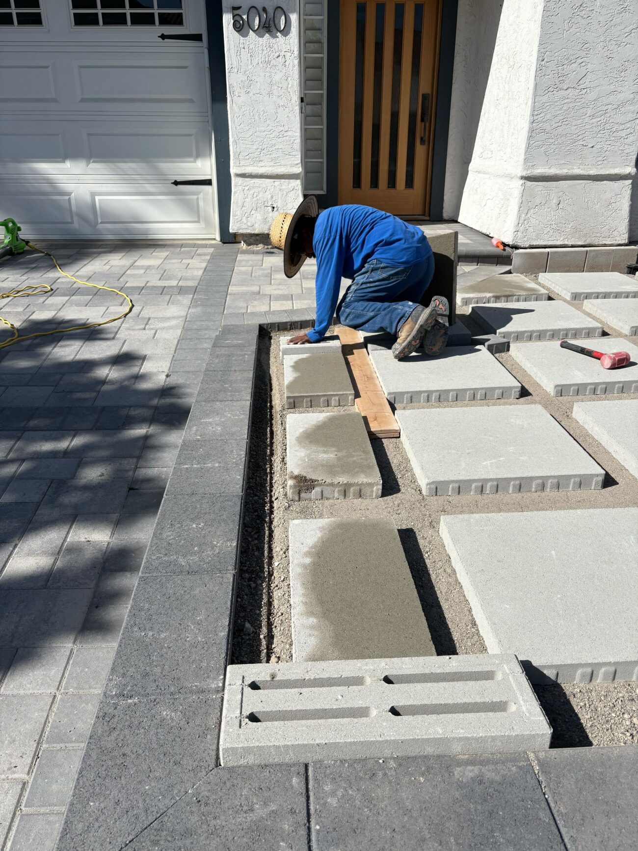 A tradesperson is kneeling and setting large concrete pavers on a compacted aggregate base at a home's front...