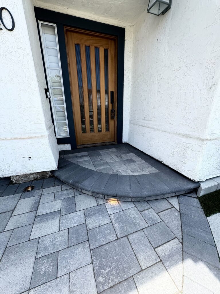 Interlocking concrete pavers in mixed gray tones form a patterned walkway and a curved entry landing leading up to a...