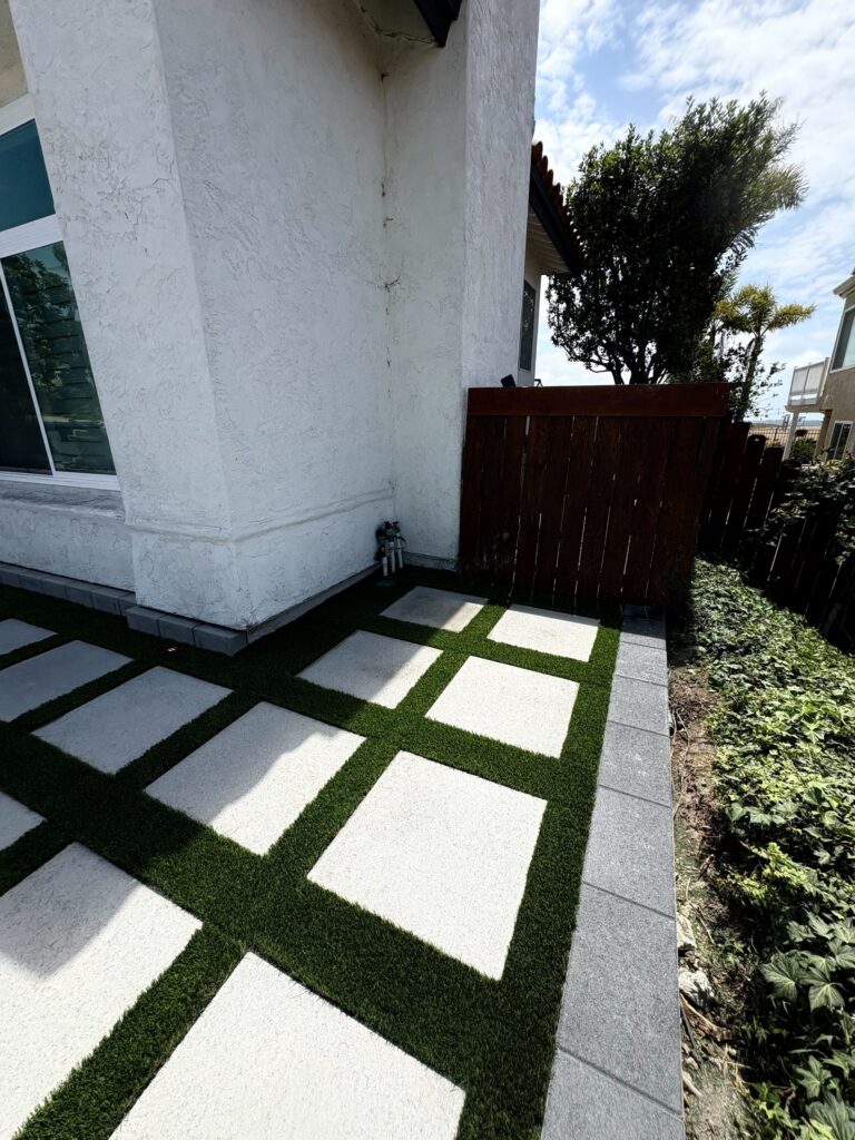 Residential side patio showing square concrete pavers laid in a checkerboard grid with artificial turf infill and a gray...