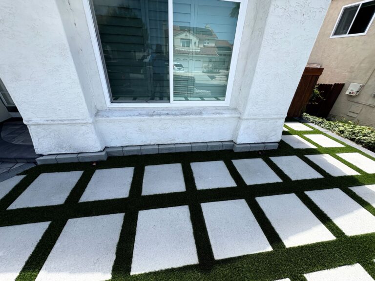 Finished residential paver patio in a checkerboard layout with large square pavers and artificial turf-filled joints...