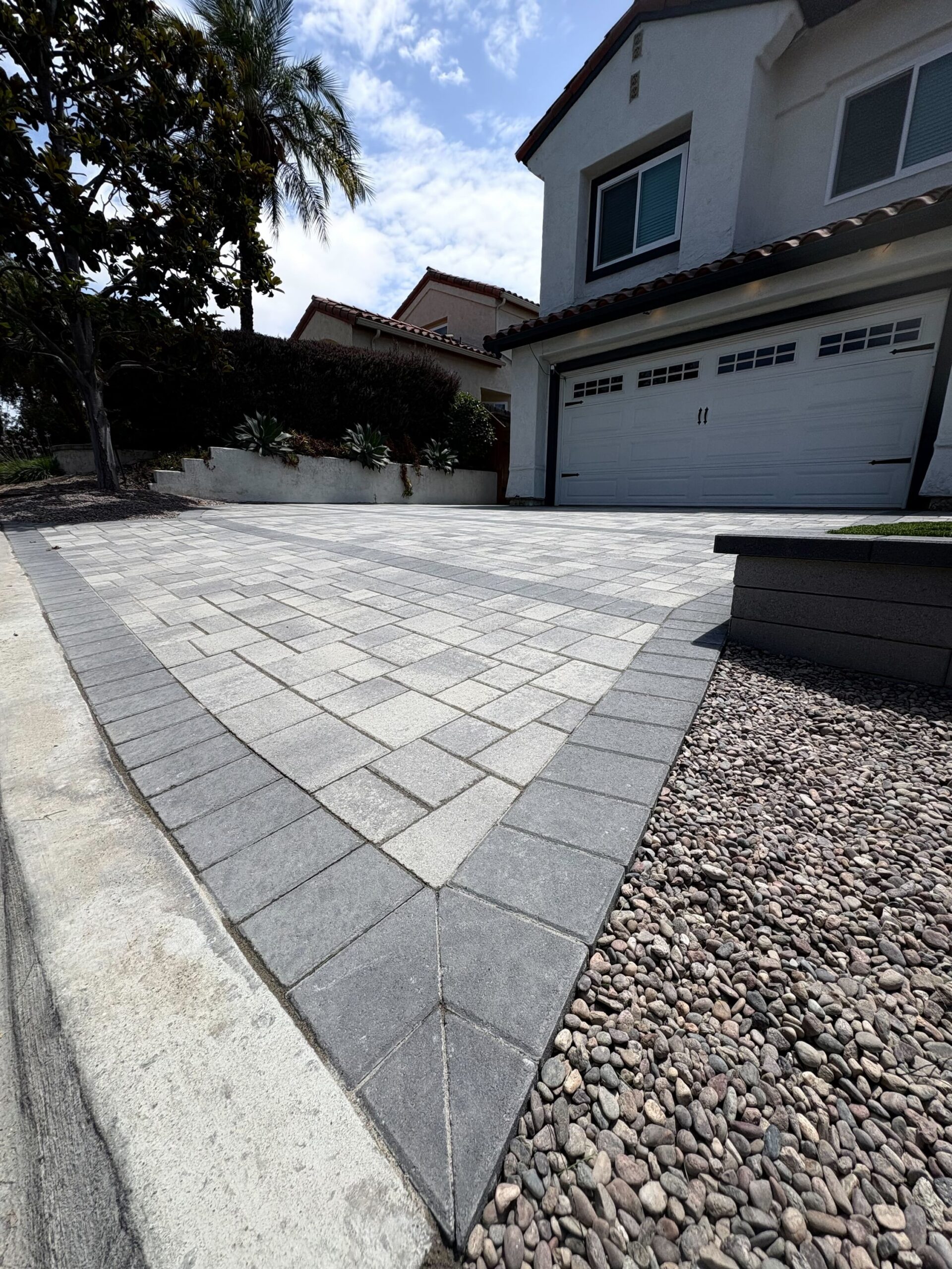 Finished gray paver driveway with a darker soldier-course border running up to the garage, river rock along one side.