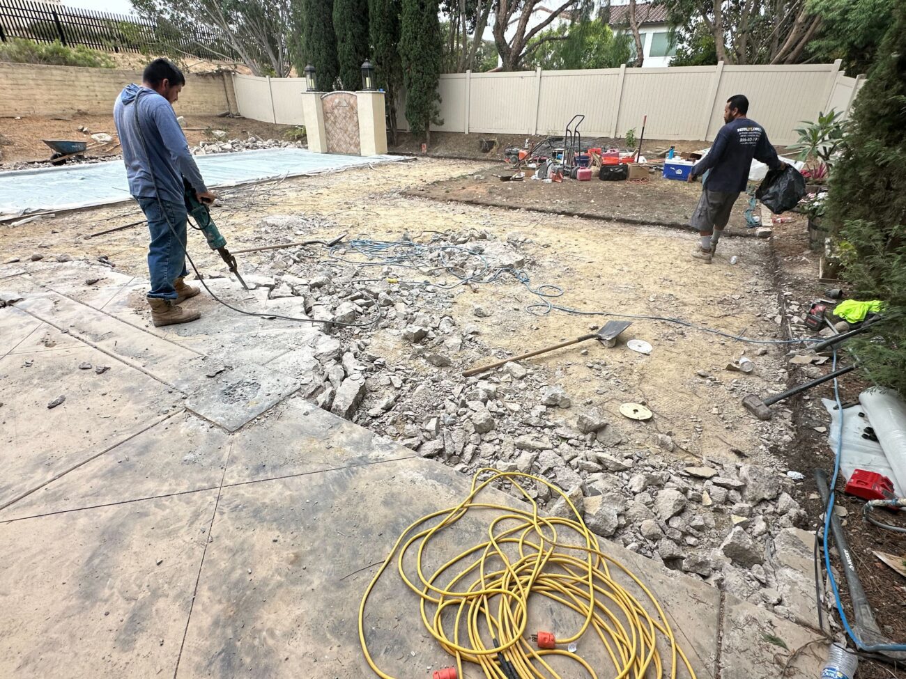 Two crew members are actively jackhammering and breaking up an existing concrete patio, leaving scattered concrete rubble...