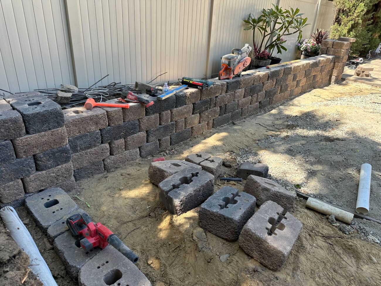 Segmental concrete block retaining wall under construction along a backyard fence. Loose blocks sit on compacted sand...