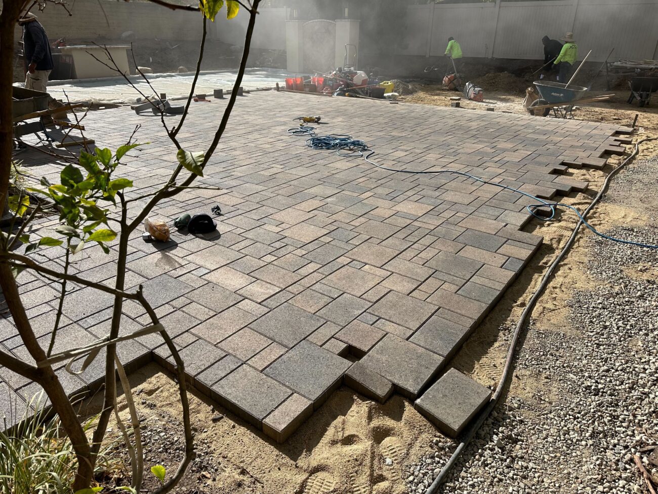 Crew actively installing interlocking pavers on a compacted sand bed to form a patio surface. The field shows a modular...
