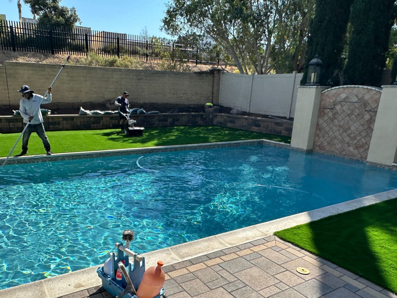Backyard pool area with crew installing artificial turf along a paver pool deck. Two workers are using turf equipment and...
